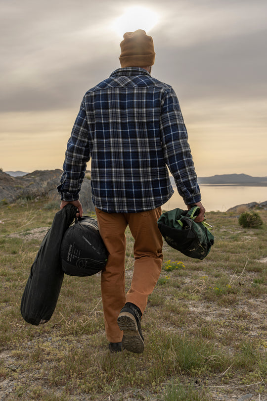 An adventurer, sporting Bulletprufe Jeans - Whiskey Denim Slim Fit, strides confidently with camping gear, ready for any ultimate outdoor experience.