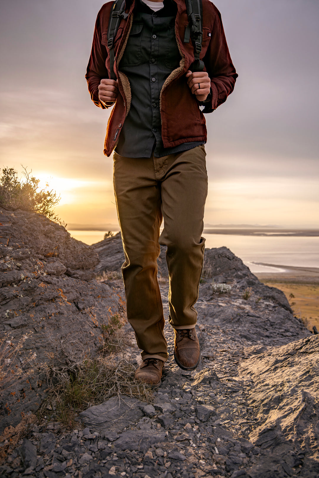 Man in Olive Green Kush Denim Jeans - Adventure Fit - 6th Gen confidently walking on a rugged rocky hill during an outdoor adventure.