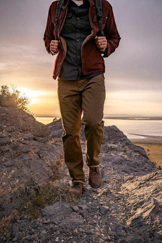 Man in Olive Green Kush Denim Jeans - Adventure Fit - 6th Gen confidently walking on a rugged rocky hill during an outdoor adventure.