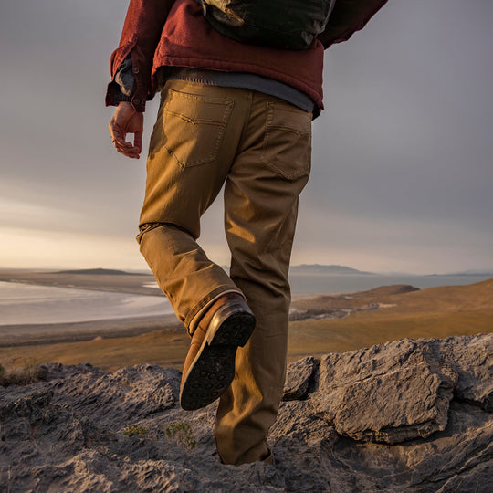 Man in Olive Green Kush Denim Jeans - Adventure Fit - 6th Gen, standing with a back view, gazing at a breathtaking outdoor horizon during an adventurous journey.
