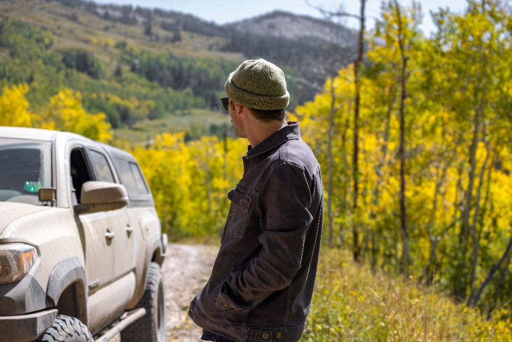 A man wearing his new Overland Jacket with the backdrop of a rugged forest. Discover its enduring design and rugged durability, ready to conquer the unknown and stand the test of time.