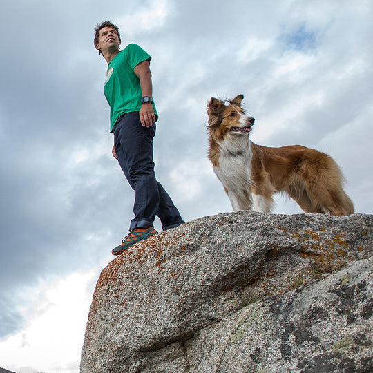 A man in Midnight Blue Denim - Slim Fit - 6th Gen stands beside a rock with his loyal dog, both immersed in the joy of an outdoor adventure.