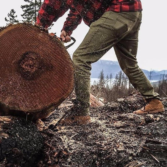 Man in Olive Green Kush Denim - Slim Fit - 6th Gen skillfully cutting a log with determination and precision.