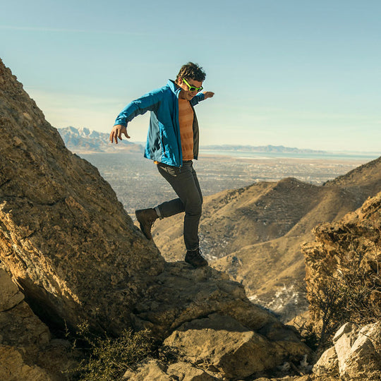 An adventurous man, dressed in Slate Grey Denim - Adventure Fit - 6th Gen, confidently navigates a precarious cliffside trail during an exhilarating outdoor excursion.