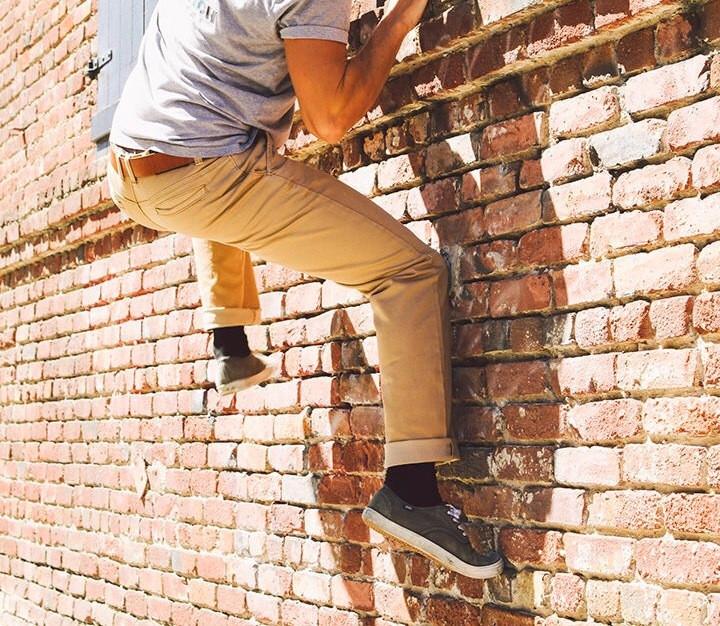 A man in Serengeti Tan Tsavo Khakis - Slim Fit Bulletprufe Jeans, scaling a wall to assess their durability for the next thrilling outdoor adventure.