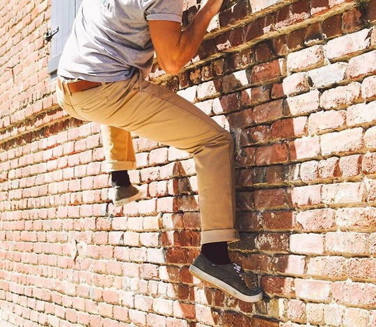 A man in Serengeti Tan Tsavo Khakis - Slim Fit Bulletprufe Jeans, scaling a wall to assess their durability for the next thrilling outdoor adventure.