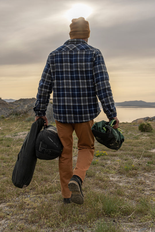 An adventurer, sporting Bulletprufe Jeans - Whiskey Denim Slim Fit, strides confidently with camping gear, ready for any ultimate outdoor experience. 
